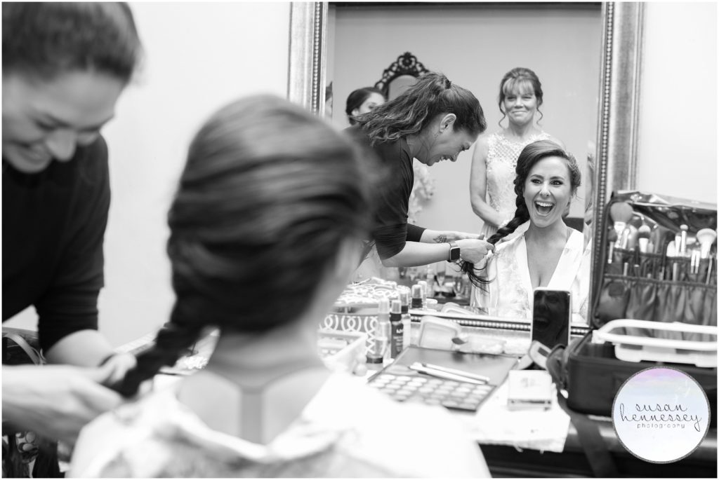 Black and white photo of a bride laughing in bridal suite while her hair is braided.